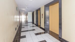Ein langer, schmaler Gang mit einem Schachbrettmuster auf dem Boden, Türen an einer Seite und einer schlichten Wand an der anderen.