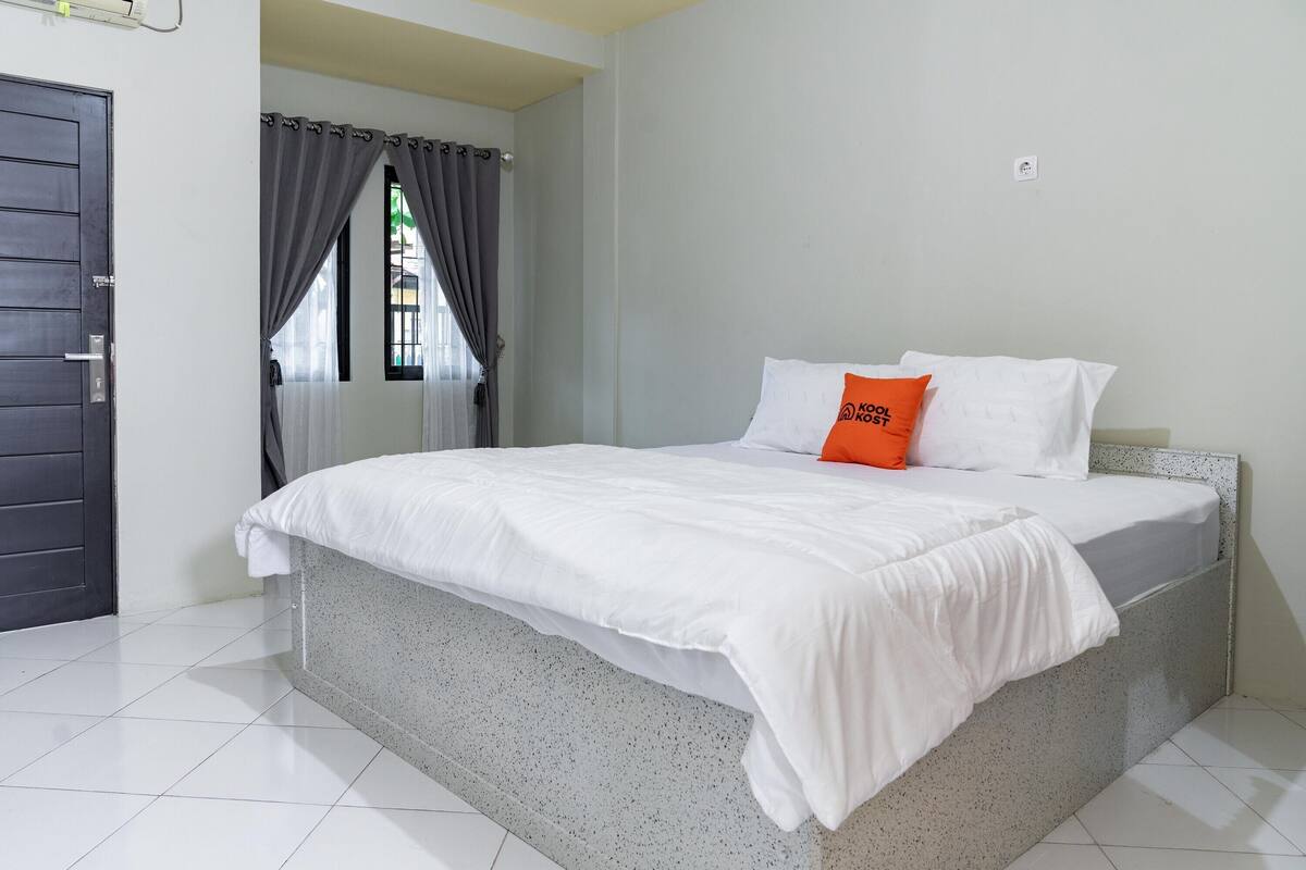 A bedroom with a bed, white bedding, a grey speckled headboard, and a window with grey curtains.