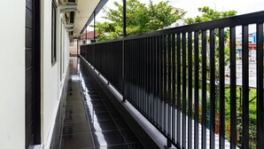 Un pasillo con barandilla de metal negro, suelo de baldosas y vistas a zonas verdes y edificios al fondo.