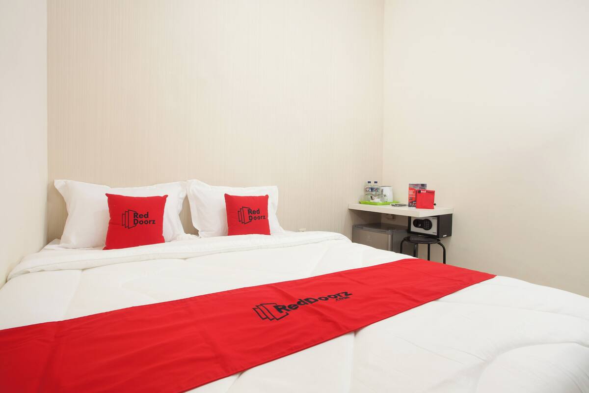 A hotel room with a neatly made bed, two pillows with a logo, a red bed runner, a small table with a radio, and a chair.