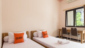 A hotel room with two single beds, each with white bedding and orange pillows, a wooden desk, two chairs, and a window with a view of greenery outside. - KoolKost Syariah near UPN Veteran (Surabaya)