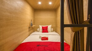 A compact room with a wooden ceiling, a bed with a red blanket, a pillow with a logo, and a small shelf with a bottle. - RedDoorz Hostel near Malang Train Station 3 (Malang)