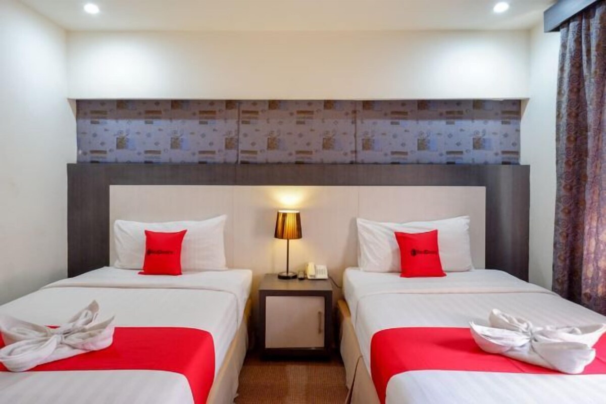 Una habitación de hotel con dos camas, cada una con una almohada roja de adorno, una mesita de noche con lámpara y un pequeño armario.