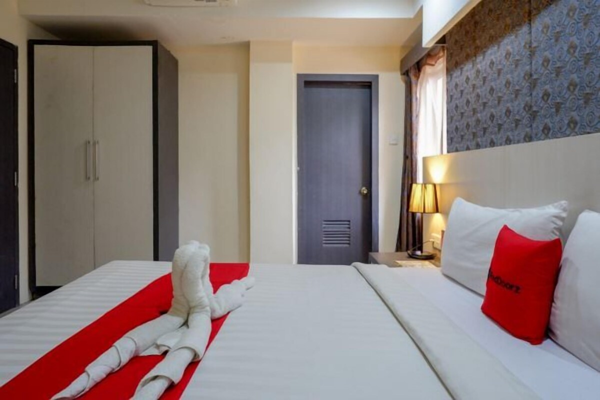 A hotel room with a bed, a red and white striped towel, a red pillow, a nightstand with a lamp, and a wardrobe.