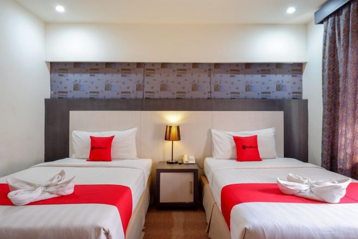 A hotel room with two beds, each with a red accent pillow, a bedside lamp, and a neatly folded towel on the beds.