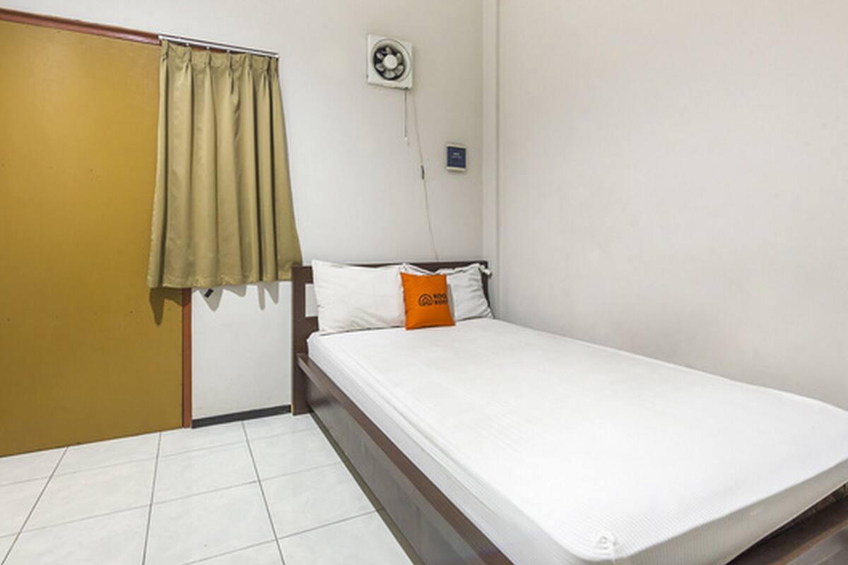A single bed with white bedding and an orange pillow, a closed door, and a wall-mounted fan.
