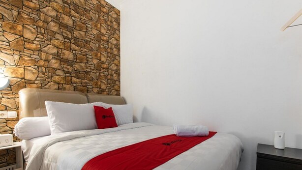 A hotel room with a stone wall, a bed with white bedding and a red throw, a nightstand with a lamp, and a dark wooden dresser. - RedDoorz Plus near Trans Studio Cibubur 2 (Bekasi)