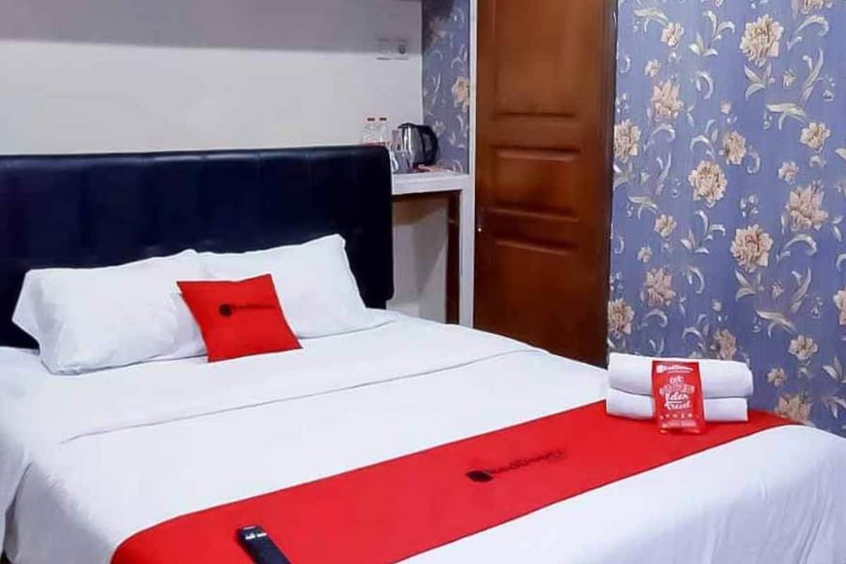 A hotel room with a bed, a red and white bedspread, a red pillow, and a red sign on the bed.