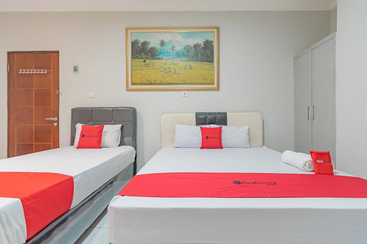 A hotel room with two beds, each with a red blanket and pillows, a painting on the wall, and a wardrobe.