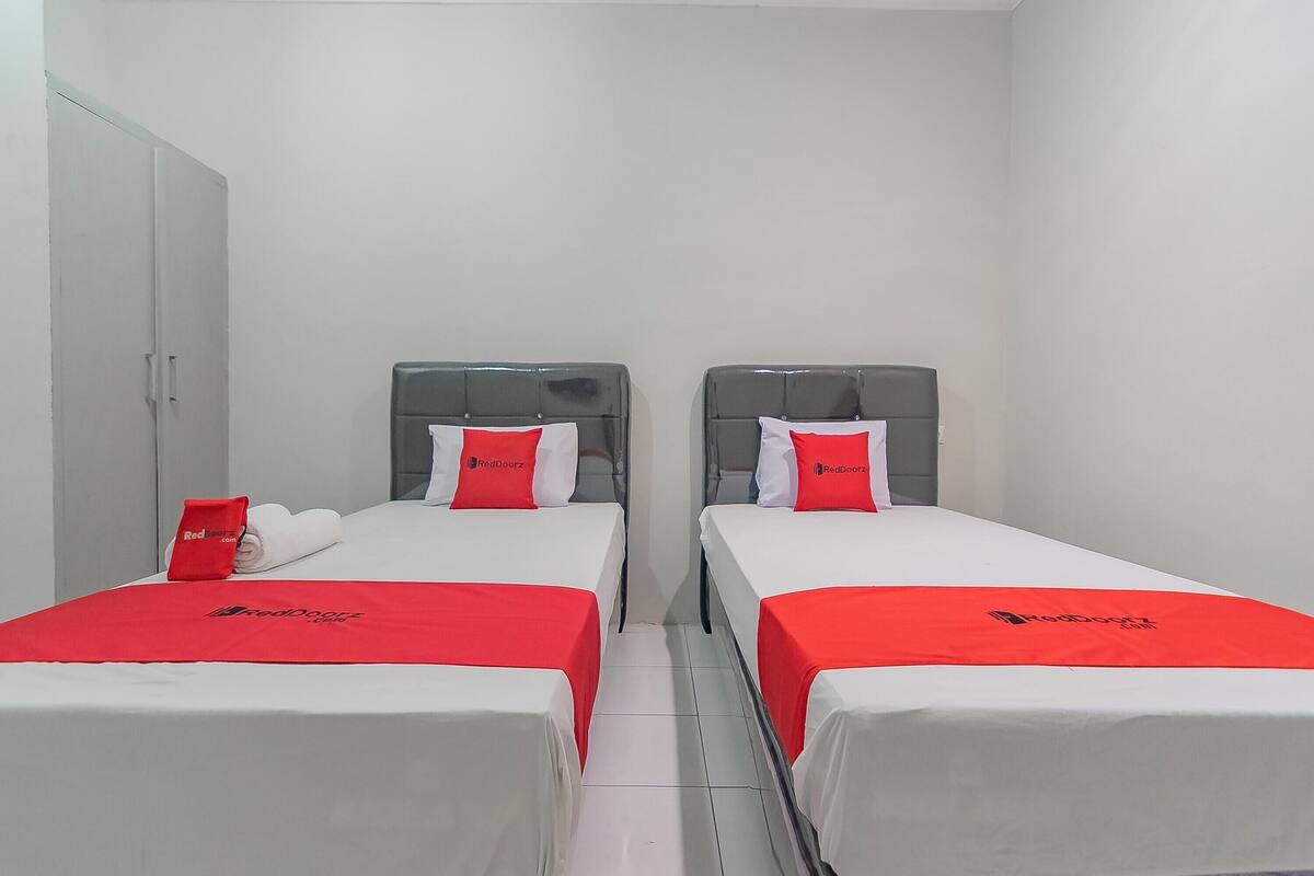 A hotel room with two single beds, each with a grey headboard and white bedding. There is a red towel and a red pillow with the hotel's logo on each bed.