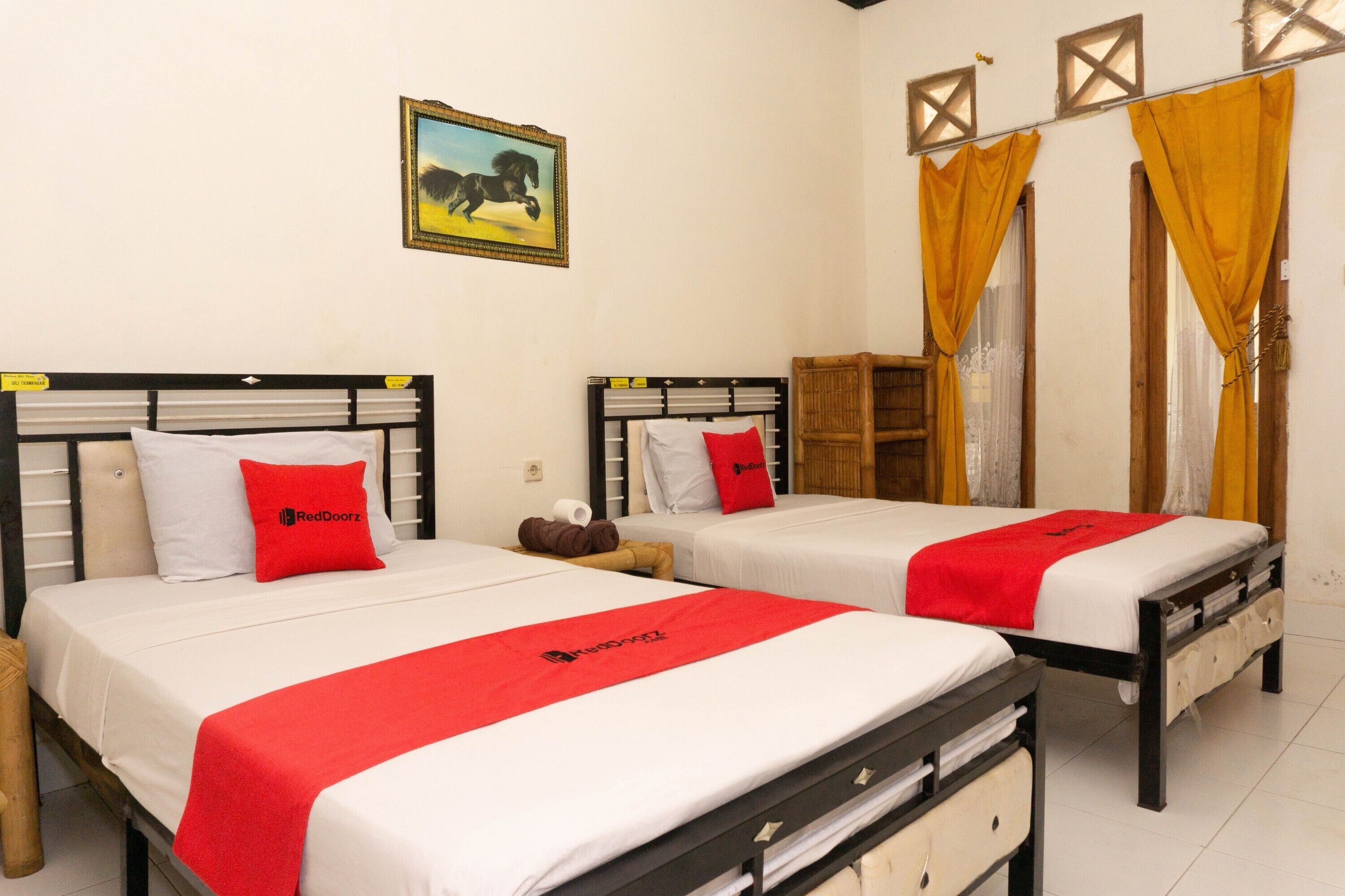 Two single beds with white bedding and red accents, a wooden wardrobe, and a painting on the wall.