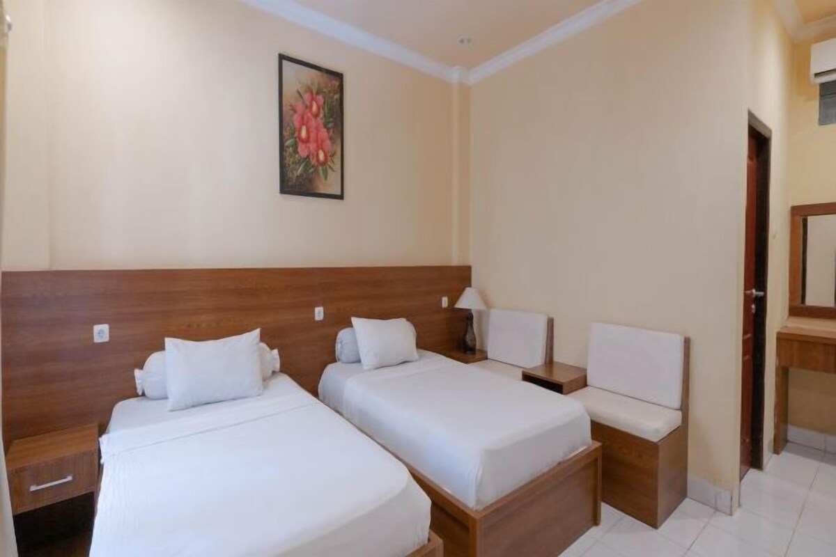 A hotel room with two single beds, a wooden headboard, a small bedside table with a lamp, and a bench.