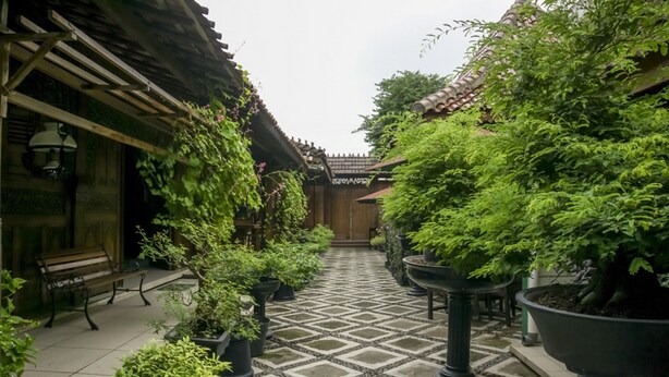 A courtyard with a tiled floor, potted plants, a wooden bench, and a traditional building with a tiled roof. - RedDoorz Syariah @ Lempuyangan (Yogyakarta)
