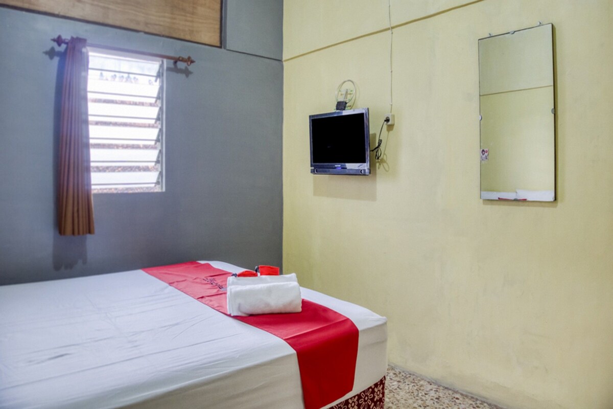 Un lit simple avec une couvre-lit rouge et blanche, une petite télévision fixée au mur et un miroir.