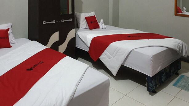 Two beds with white bedding and red accents, a dark door, and a small mirror on the wall. - RedDoorz @ Hotel Yaki Mamuju (Mamuju)