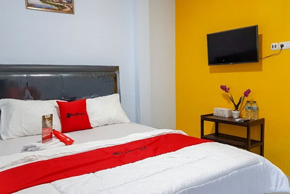 Habitación de hotel con una cama, una colcha roja y blanca, un almohadón rojo, una mesita con un radio y botellas, y un televisor montado en una pared amarilla.