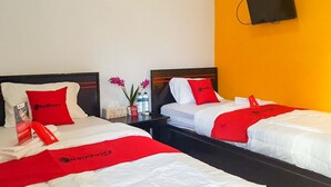 Habitación de hotel con dos camas, cada una con cubrecama rojo y blanco, un televisor de pantalla plana montado en la pared y una mesita con un jarrón de flores.