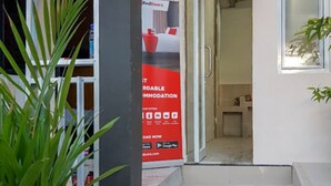 Entrada de hotel con un anuncio rojo de alojamiento económico, una planta en maceta y una vista del interior.