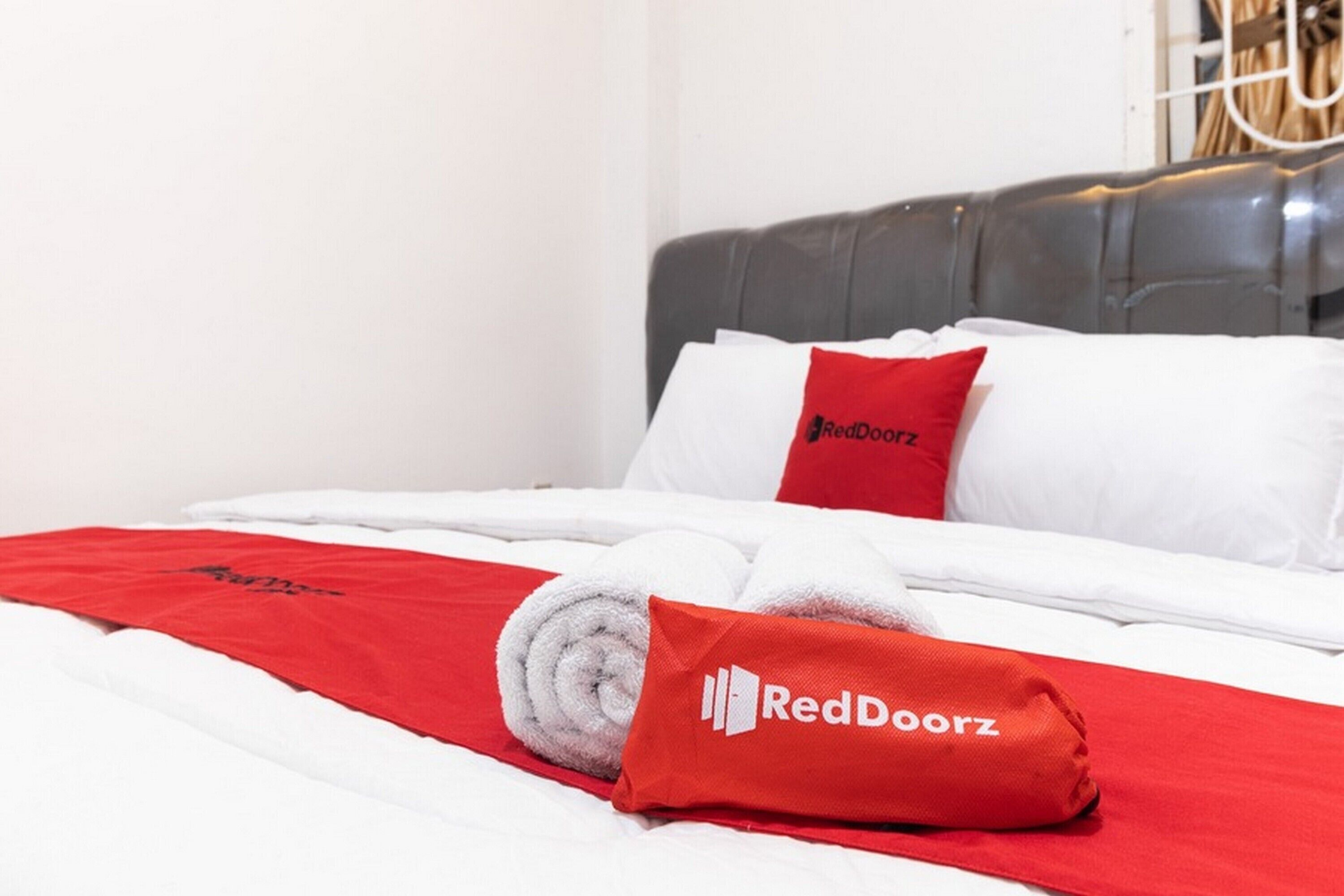 Un lit bien fait, avec des draps blancs, une couverture rouge et des oreillers ornés du logo RedDoorz.