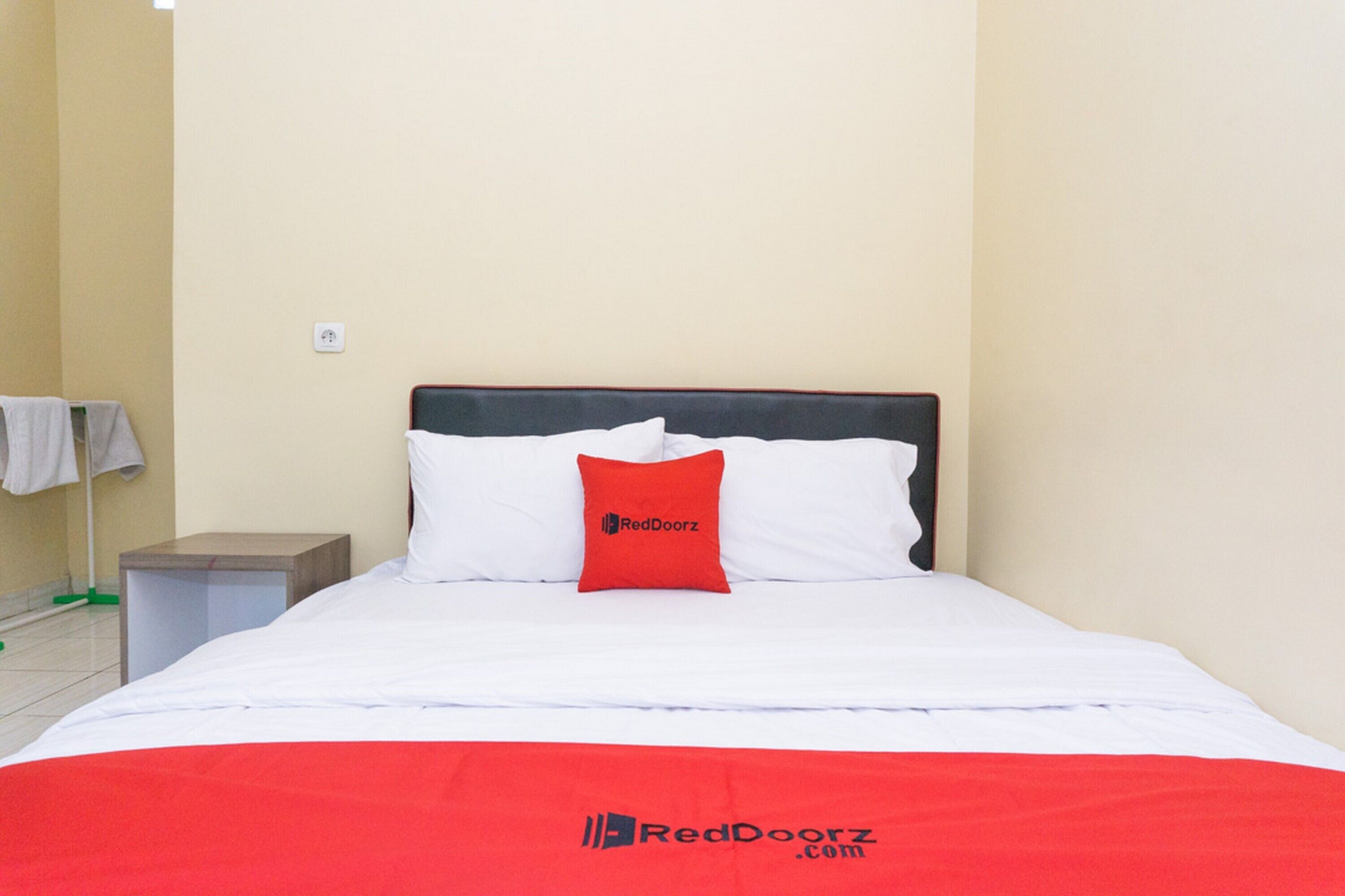 A neatly made bed with white linens and a red pillow, a small bedside table with a lamp, and a wall-mounted towel rack.