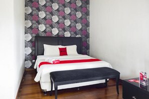 Un lit bien fait, avec une literie blanche et un coussin rouge, un banc noir et un papier peint à motifs floraux.