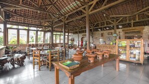 A spacious indoor area with wooden tables and chairs, a large open shelf with pottery, and a brick wall with a clock. - RedDoorz Plus @ Purwodadi (Lawang)