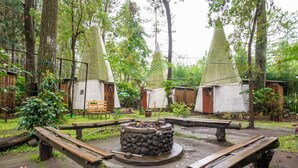 Un cadre extérieur rustique comprenant trois cabanes à toit conique, un foyer central en pierre et des bancs en bois.