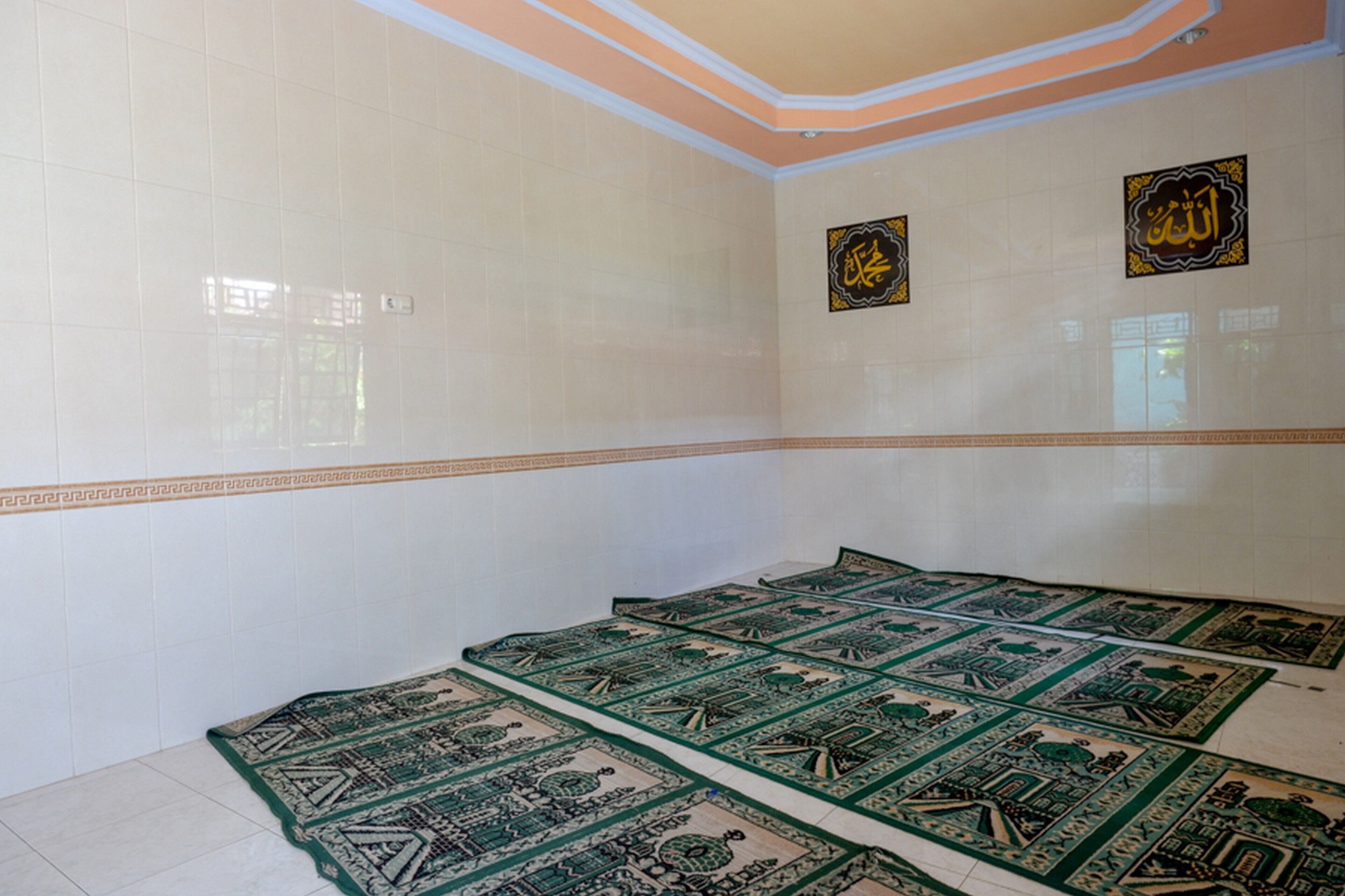 A room with white tiled walls, a green patterned carpet, and Arabic calligraphy on the wall.