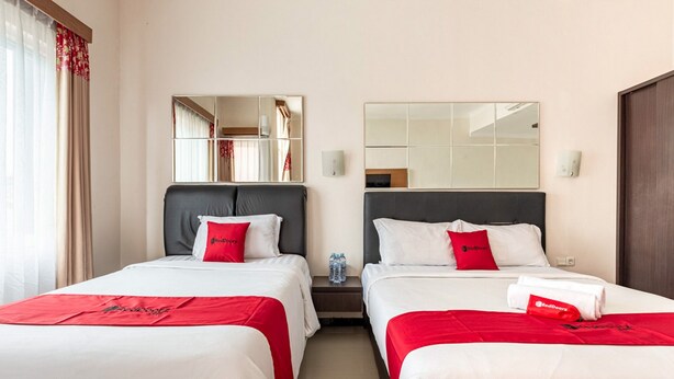 Une chambre d’hôtel avec deux lits, chacun recouvert d’une couverture rouge et blanche, une table de chevet avec une bouteille d’eau et un miroir au-dessus de chaque lit.