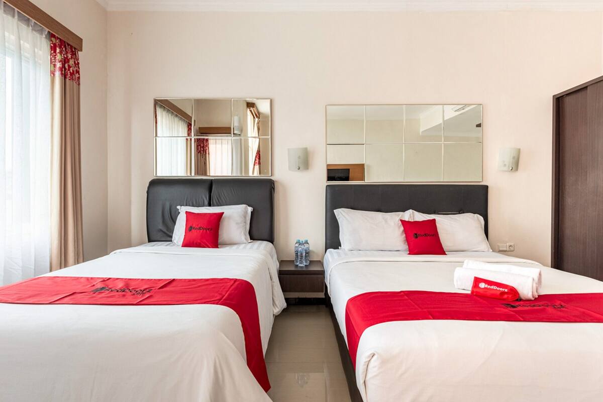 Habitación de hotel con dos camas, cada una con cubrecama rojo y blanco, una mesita de noche con botella de agua y un espejo sobre cada cama.