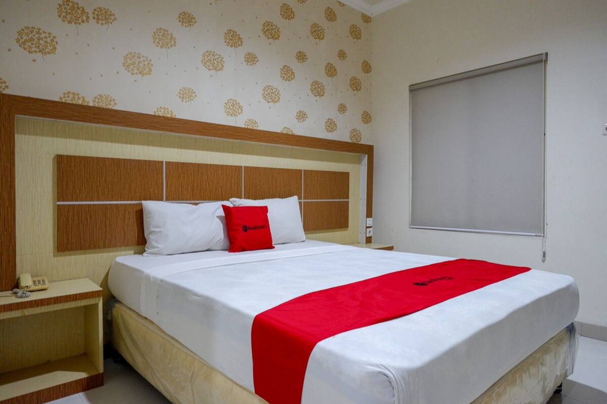 Una habitación de hotel con una cama grande, cabecera de madera, manta roja y papel tapiz estampado.