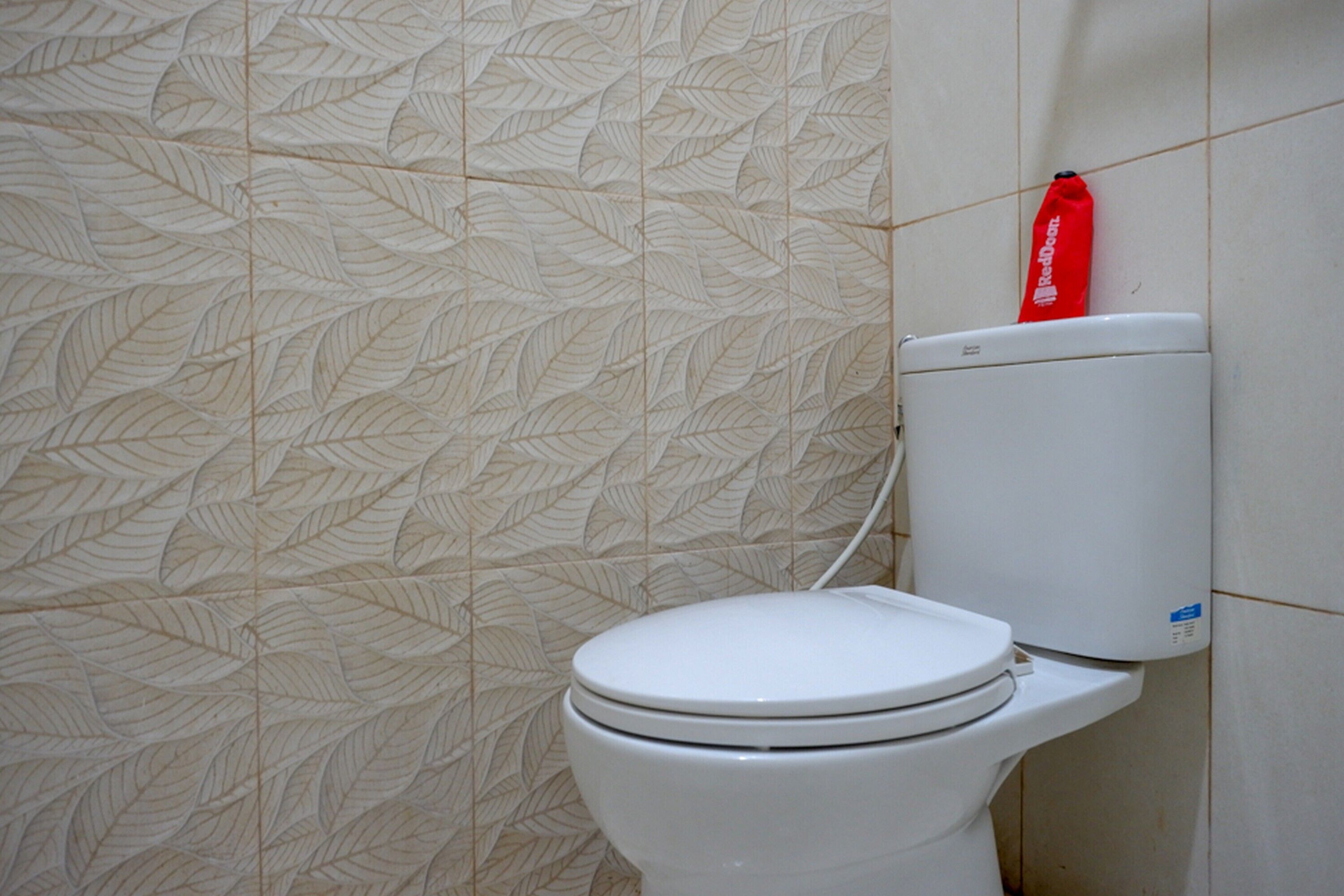 Eine weiße Toilette mit einem roten Müllbeutel darauf, vor einer Wand mit einem Blattmuster.