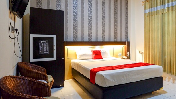 A hotel room with a bed, a television mounted on the wall, a chair, and a window with curtains. - RedDoorz @ Hotel Bumi Palupy (Palu)