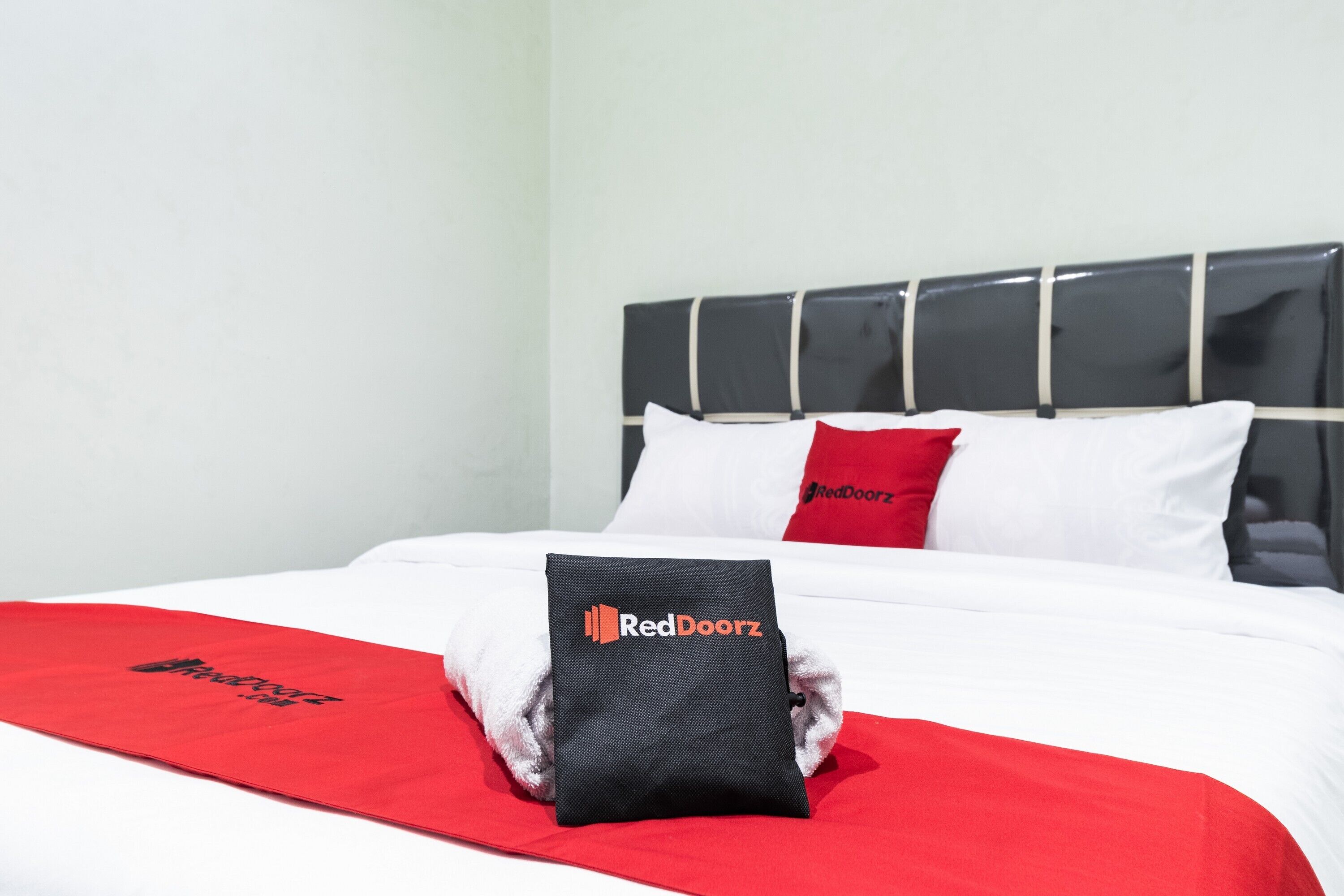 A neatly made bed with a red and white bedspread, a black pillow with the RedDoorz logo, and a red pillow.