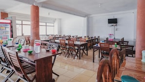 A spacious dining area with multiple wooden tables and chairs, a refrigerator with beverages, and a television mounted on the wall. - RedDoorz Syariah near Kebun Raya Liwa (Gunung Sugih)