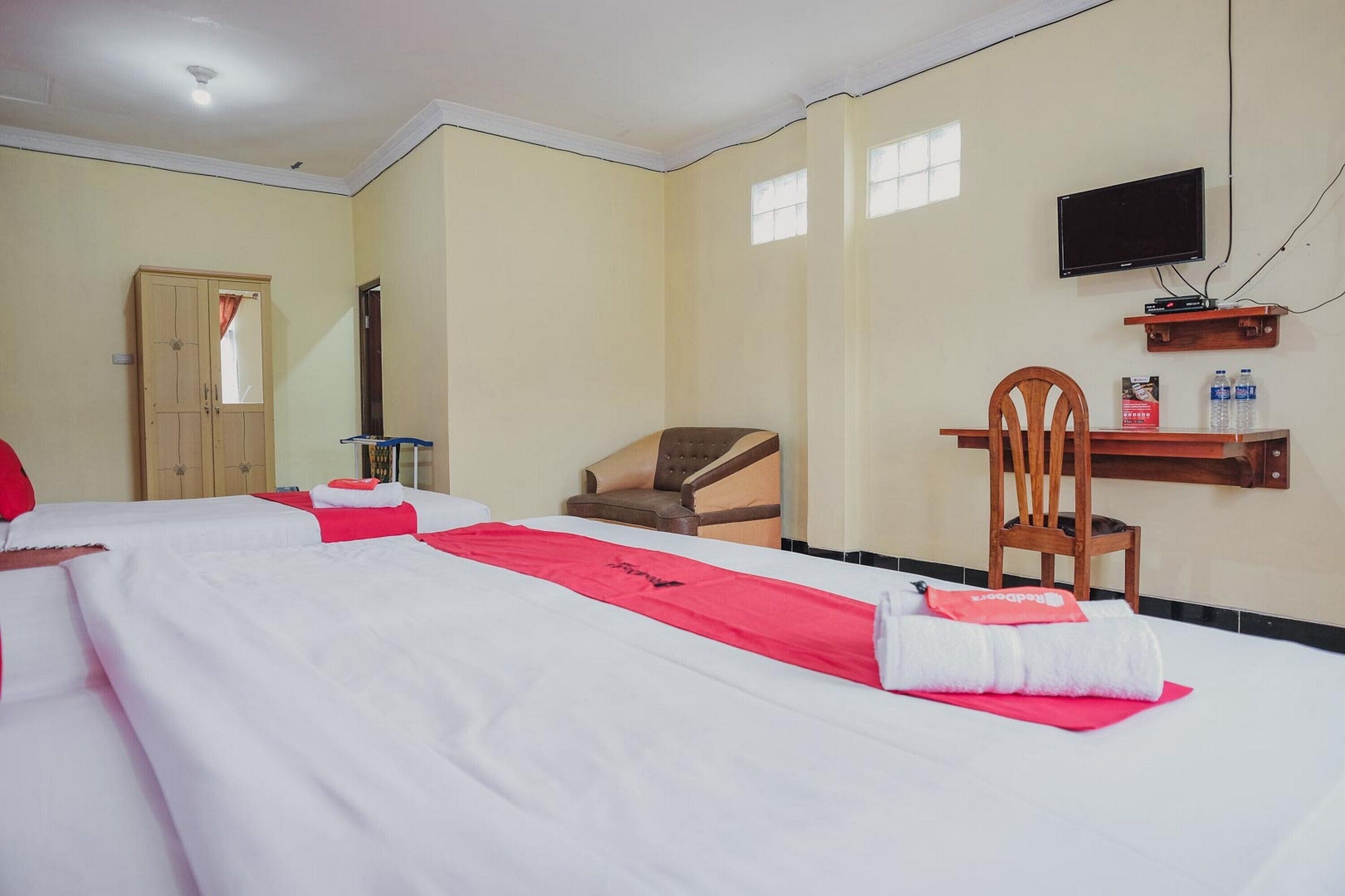 A hotel room with two beds, a TV, a wooden chair, and a shelf with bottled water.