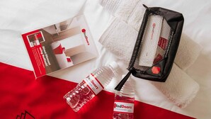 Une bouteille d’eau, une serviette et une boîte, tous aux couleurs de la marque RedDoorz, posés sur un couvre-lit rouge et blanc.