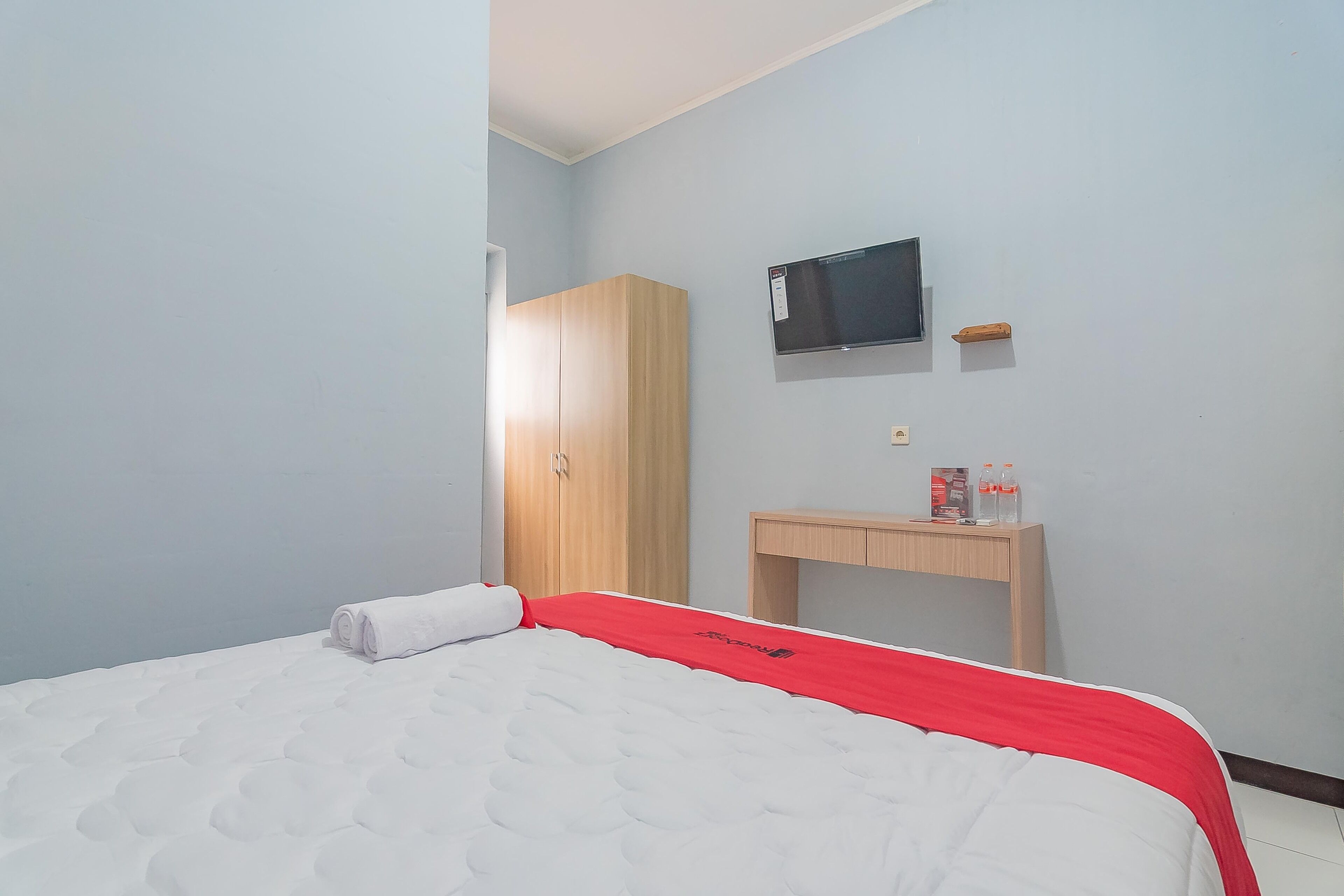 A hotel room with a bed, a wooden wardrobe, a TV mounted on the wall, and a small table with a bottle and a box on it.