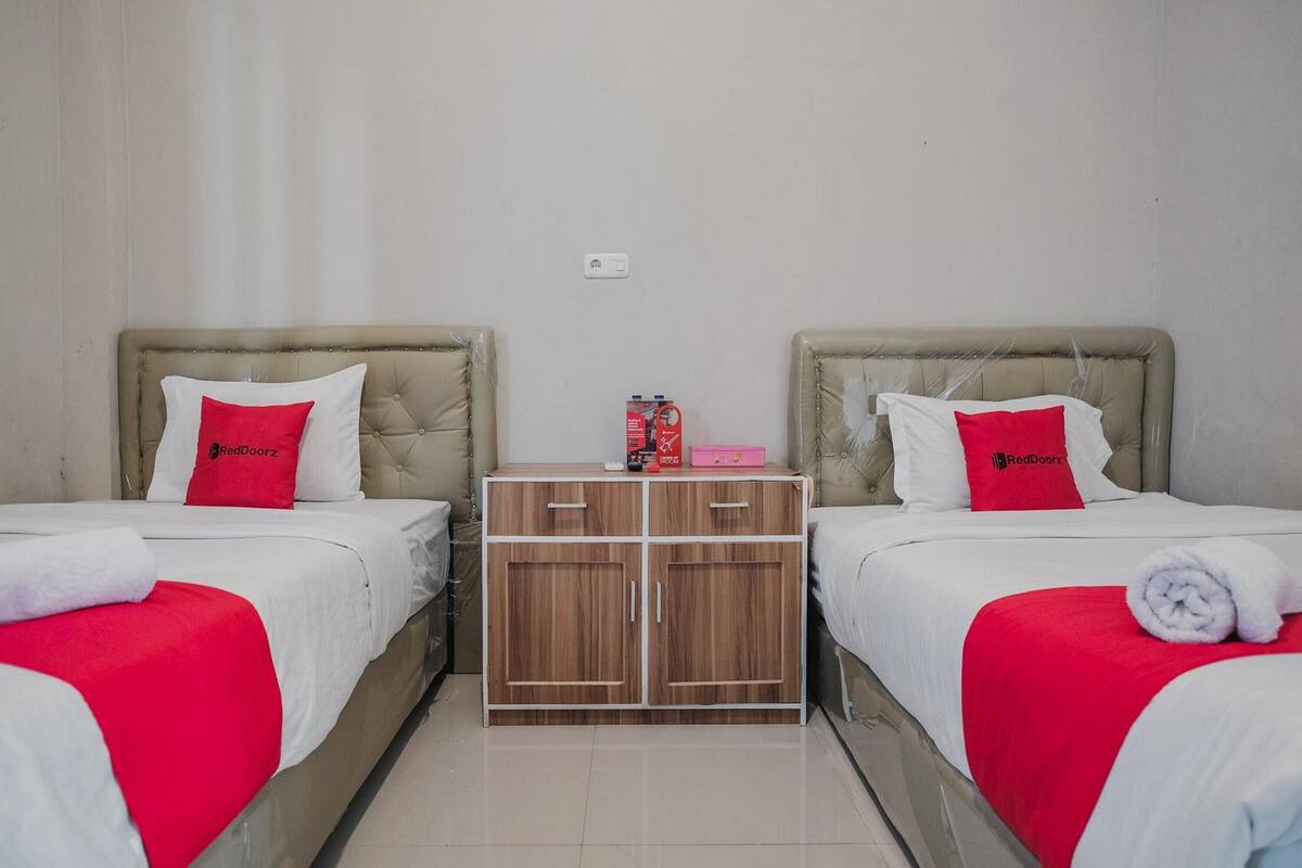A hotel room with two beds, each with a red pillow and white bedding. There is a wooden wardrobe with a mirror and a small table with a red box and a pink item on it.