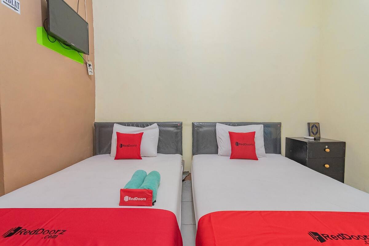 A hotel room with two beds, each with a red blanket and pillow, and a small bedside table with a lamp.