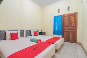 Habitación de hotel con dos camas, cada una con una colcha roja y un cojín con el logotipo del hotel. Se observa una puerta de madera y una mesita de noche con una lámpara.