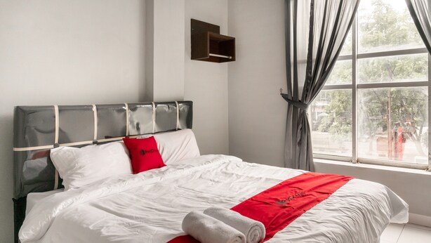 Una cama bien hecha con ropa de cama blanca y roja, cabecera y ventana con cortinas.