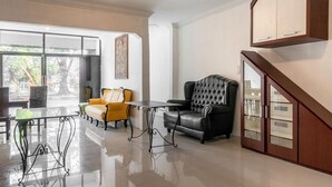 A spacious living area with a black leather sofa, a yellow armchair, and a glass-top coffee table. A wooden cabinet is mounted on the wall, and a staircase is visible to the right. - RedDoorz near Manhattan Times Square Medan 2 (Medan)