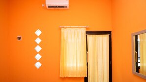 A bathroom with an orange wall, a window with yellow curtains, and a white air conditioner mounted above the window. - KoolKost Syariah near Transmart Tegal (Tegal)