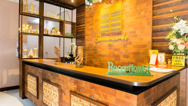 A reception desk with a wooden finish, a sign reading "Samwombo Hotel", and a "Receptionist" sign. There are decorative items on shelves and a "Free WiFi Area" sign.