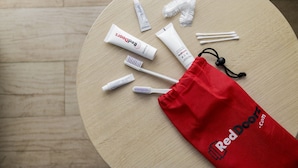Pyöreä puinen pöytä, jonka päällä on punainen nyörikuorinen pussi, jossa on merkintä ”RedDoorz.com”. Pussin sisällä on suuhygieniatuotteita, kuten hammastahnaa, hammasharja ja hammaslankaa.