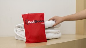 Käsi asettaa punaisen, tekstillä ”Red Doorz.com” varustetun kassin valkoisten pyyhkeiden päälle puupinnalle.