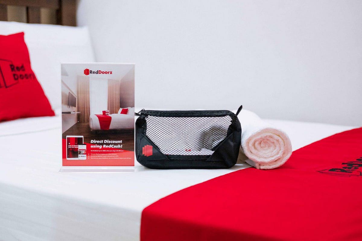 A hotel room with a bed, a RedDoorz branded box, and a black mesh bag with a rolled-up towel.