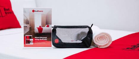 Una habitación de hotel con una cama bien hecha, un folleto promocional de RedDoorz, una bolsa de malla negra y una almohada roja.