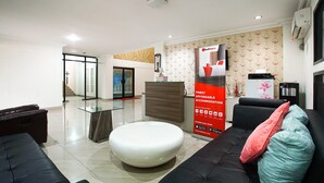 A modern living room with a sectional sofa, a round ottoman, and a glass coffee table. There is a reception desk with a RedDoorz sign and a large poster advertising accommodation. - RedDoorz near Palembang Trade Center 3 (Palembang)